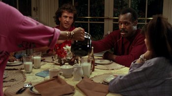 Movie still from “Lethal Weapon” (1987), directed by Richard Donner – A group of people sitting at a table with food; Medium shot, Over the shoulder angle