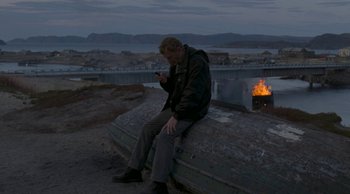 Movie still from “Leviathan” (2014), directed by Andrey Zvyagintsev – A man sitting on top of a rock near a fire pit; Wide shot, High angle