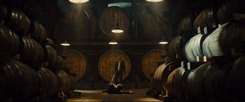 Movie still from “Kingsman: The Golden Circle” (2017), directed by Matthew Vaughn – A man in a suit and a hat laying on the ground in front of a bunch of wooden barrels; Wide shot, Low angle