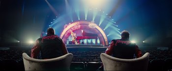 Movie still from “Kingsman: The Golden Circle” (2017), directed by Matthew Vaughn – Two people sitting in chairs in front of a stage with a piano; Wide shot, Low angle