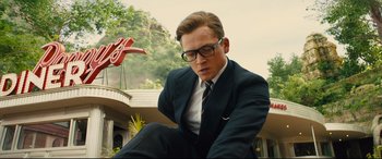 Movie still from “Kingsman: The Golden Circle” (2017), directed by Matthew Vaughn – A man in a suit and tie sitting in front of a building; Medium shot, Low angle