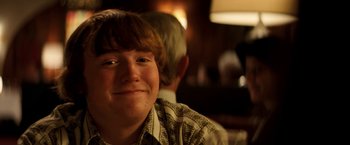 Movie still from “Licorice Pizza” (2021), directed by Paul Thomas Anderson – A young boy smiling for the camera with his father in the background; Close Up shot, Low angle