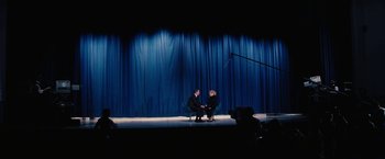 Movie still from “Licorice Pizza” (2021), directed by Paul Thomas Anderson – Two people are sitting on a stage with a curtain behind them; Extreme Wide shot, High angle