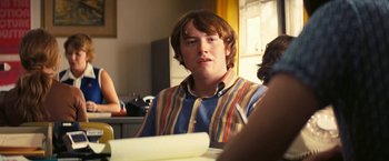 Movie still from “Licorice Pizza” (2021), directed by Paul Thomas Anderson – A man sitting in front of a table with papers in front of him; Medium shot, Over the shoulder angle