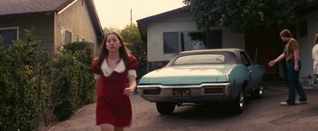 Movie still from “Licorice Pizza” (2021), directed by Paul Thomas Anderson – A woman in a red dress walking past an old car; Wide shot, High angle
