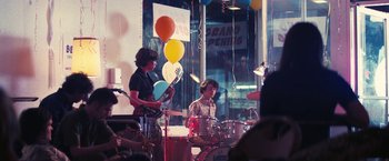 Movie still from “Licorice Pizza” (2021), directed by Paul Thomas Anderson – A group of young men playing musical instruments in front of balloons; Wide shot, Over the shoulder angle