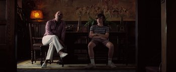 Movie still from “Licorice Pizza” (2021), directed by Paul Thomas Anderson – Two men sitting on a chair in front of a painting; Medium shot, Low angle