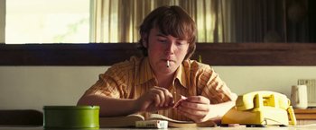 Movie still from “Licorice Pizza” (2021), directed by Paul Thomas Anderson – A young man sitting at a table with a cigarette in his mouth; Medium shot, Low angle