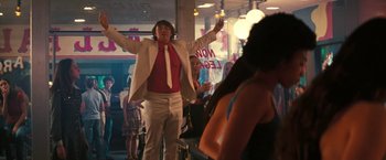 Movie still from “Licorice Pizza” (2021), directed by Paul Thomas Anderson – A man in a white jacket and a red tie is holding his arms up; Medium shot, Over the shoulder angle