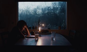 Movie still from “Light of My Life” (2019), directed by Casey Affleck – A person sitting at a table with a candle in a dark room; Medium shot, Over the shoulder angle