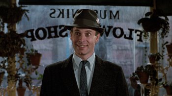 Movie still from “Little Shop of Horrors” (1986), directed by Frank Oz – A man wearing a suit and a hat smiling for the camera; Close Up shot, Over the shoulder angle