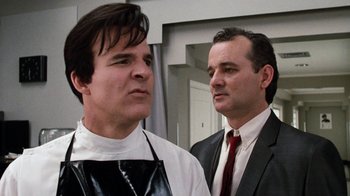 Movie still from “Little Shop of Horrors” (1986), directed by Frank Oz – A man in an apron standing next to another man in a suit and tie; Close Up shot, Low angle