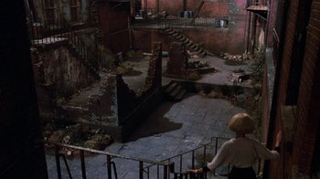 Movie still from “Little Shop of Horrors” (1986), directed by Frank Oz – A woman standing in a room that has a lot of stairs in it; Extreme Wide shot, High angle