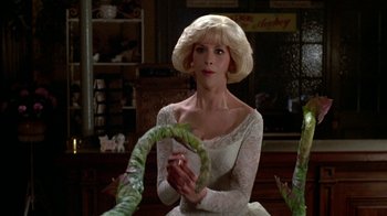Movie still from “Little Shop of Horrors” (1986), directed by Frank Oz – A woman in a white dress holding a green plant; Medium shot, Over the shoulder angle