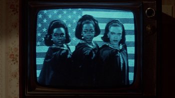 Movie still from “Little Shop of Horrors” (1986), directed by Frank Oz – A television screen showing three women in front of an american flag; Medium shot, Low angle