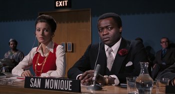 Movie still from “Live and Let Die” (1973), directed by Guy Hamilton – A man and a woman are sitting at a table with microphones; Medium shot, Over the shoulder angle