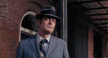Movie still from “Live and Let Die” (1973), directed by Guy Hamilton – A man in a suit and hat standing in front of a building; Close Up shot, Low angle