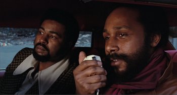 Movie still from “Live and Let Die” (1973), directed by Guy Hamilton – Two men sitting in the back of a car holding a game controller; Close Up shot, Low angle