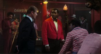 Movie still from “Live and Let Die” (1973), directed by Guy Hamilton – A group of men standing in a room; Medium shot, Over the shoulder angle