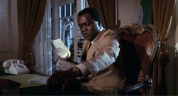 Movie still from “Live and Let Die” (1973), directed by Guy Hamilton – A man sitting in a chair holding a paper; Medium shot, Low angle