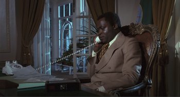 Movie still from “Live and Let Die” (1973), directed by Guy Hamilton – A man sitting in a chair talking on a phone; Medium shot, Low angle