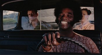 Movie still from “Live and Let Die” (1973), directed by Guy Hamilton – A man driving a car while another man watches from the passenger seat; Medium shot, Low angle