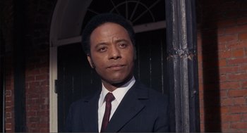 Movie still from “Live and Let Die” (1973), directed by Guy Hamilton – A man wearing a suit and tie standing in front of a building; Close Up shot, Low angle