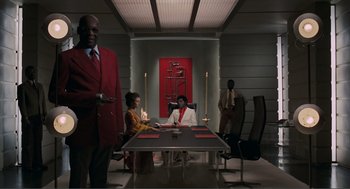 Movie still from “Live and Let Die” (1973), directed by Guy Hamilton – A group of people sitting at a table in front of a painting; Wide shot, Low angle