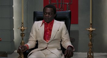 Movie still from “Live and Let Die” (1973), directed by Guy Hamilton – A man sitting in a chair wearing a white suit and red shirt; Medium shot, Low angle
