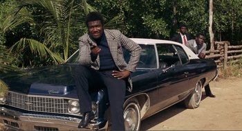 Movie still from “Live and Let Die” (1973), directed by Guy Hamilton – A man sitting on the hood of a car; Medium shot, Low angle