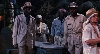Movie still from “Live and Let Die” (1973), directed by Guy Hamilton – A group of people standing around a table; Wide shot, High angle