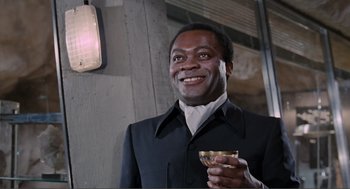 Movie still from “Live and Let Die” (1973), directed by Guy Hamilton – A man holding a wine glass in his right hand; Medium shot, Low angle