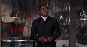 Movie still from “Live and Let Die” (1973), directed by Guy Hamilton – A man in a black suit standing next to a woman in a red shirt; Medium shot, Low angle