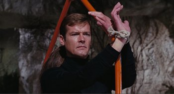 Movie still from “Live and Let Die” (1973), directed by Guy Hamilton – A man in a black shirt is chained to a metal pole; Close Up shot, High angle