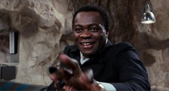 Movie still from “Live and Let Die” (1973), directed by Guy Hamilton – A man in a black suit is clapping; Close Up shot, Low angle