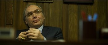 Movie still from “Logan Lucky” (2017), directed by Steven Soderbergh – A man in a suit and glasses sitting at a table; Close Up shot, Over the shoulder angle