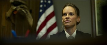 Movie still from “Logan Lucky” (2017), directed by Steven Soderbergh – A woman wearing a suit and tie in front of an american flag; Close Up shot, Low angle