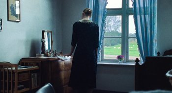 Movie still from “Lore” (2012), directed by Cate Shortland – A woman standing in front of a window looking out at a field; Medium shot, Over the shoulder angle