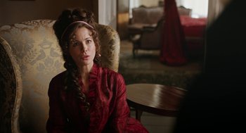 Movie still from “Love & Friendship” (2016), directed by Whit Stillman – A woman in a red dress sitting in a chair in a room; Medium shot, Over the shoulder angle