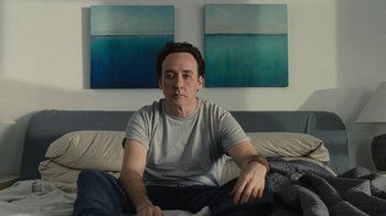Movie still from “Love & Mercy” (2014), directed by Bill Pohlad – A man sitting on top of a bed next to two paintings on the wall; Medium shot, Low angle