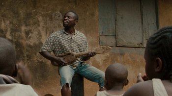 Movie still from “Machine Gun Preacher” (2011), directed by Marc Forster – A man sitting on the side of a building playing a guitar; Medium shot, Low angle