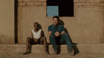 Movie still from “Machine Gun Preacher” (2011), directed by Marc Forster – A man sitting next to a young boy in front of a brick wall; Wide shot, Low angle