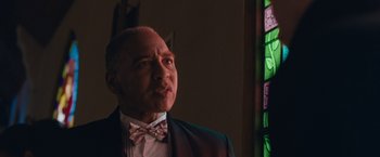 Movie still from “Little Fish” (2020), directed by Chad Hartigan – A man wearing a suit and a bow tie in front of a stained glass window; Close Up shot, Low angle