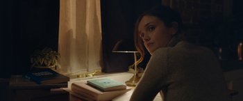 Movie still from “Little Fish” (2020), directed by Chad Hartigan – A woman sitting at a table with a book and a lamp; Medium shot, Over the shoulder angle