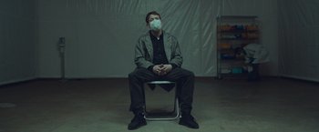 Movie still from “Little Fish” (2020), directed by Chad Hartigan – A man sitting on top of a chair wearing a face mask; Medium shot, High angle