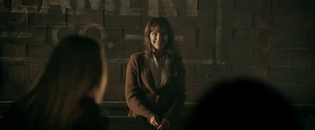 Movie still from “Malignant” (2021), directed by James Wan – A woman sitting in front of a brick wall smiling; Medium shot, Over the shoulder angle