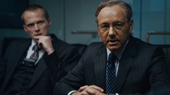 Movie still from “Margin Call” (2011), directed by J.C. Chandor – A man in a suit sitting next to another man in a suit; Close Up shot, Over the shoulder angle