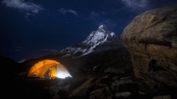 A tent is lit up in the night sky near a mountain peak