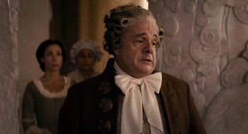 Movie still from “Mirror Mirror” (2012), directed by Tarsem Singh – An older man with a wig and a bow tie; Medium shot, Over the shoulder angle