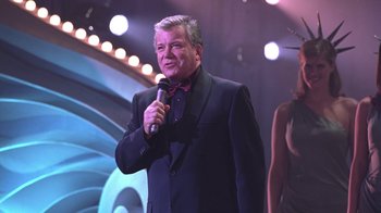 Movie still from “Miss Congeniality” (2000), directed by Donald Petrie – A man holding a microphone while standing in front of an audience; Medium shot, Over the shoulder angle